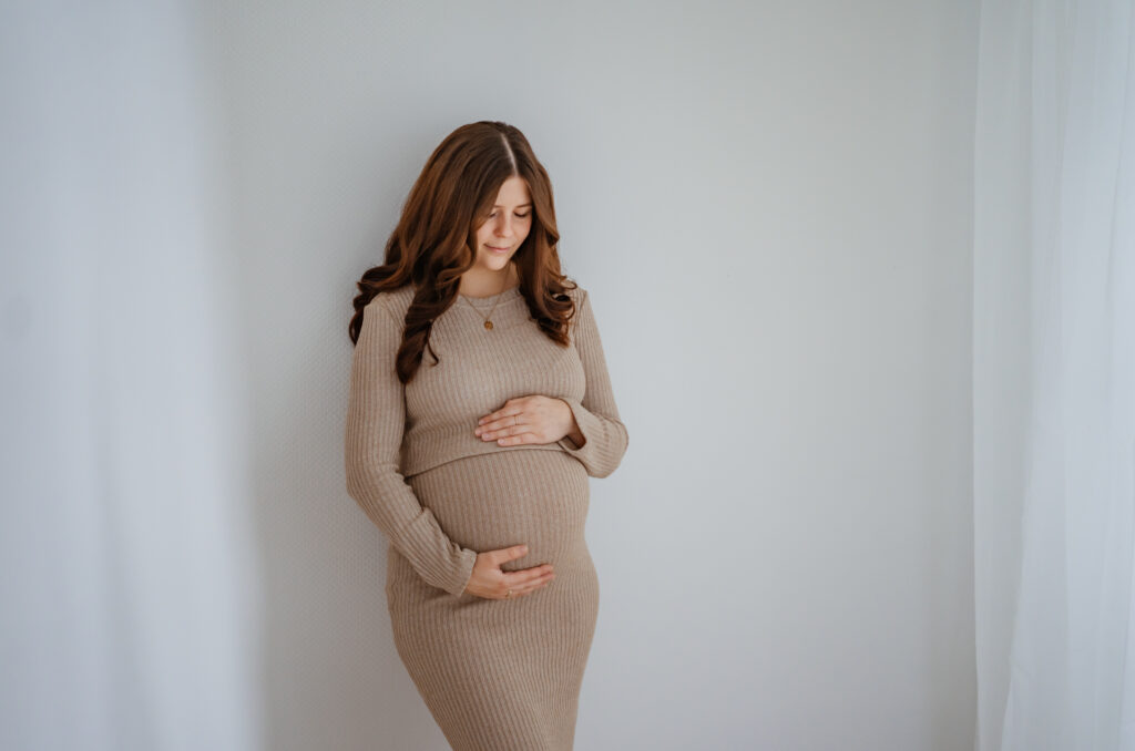 Ein schlichtes Bild bei dem eine werdende Mutter ihren Babybauch hält und an der Wand im Tageslichtatelier in Kreuzwertheim lehnt