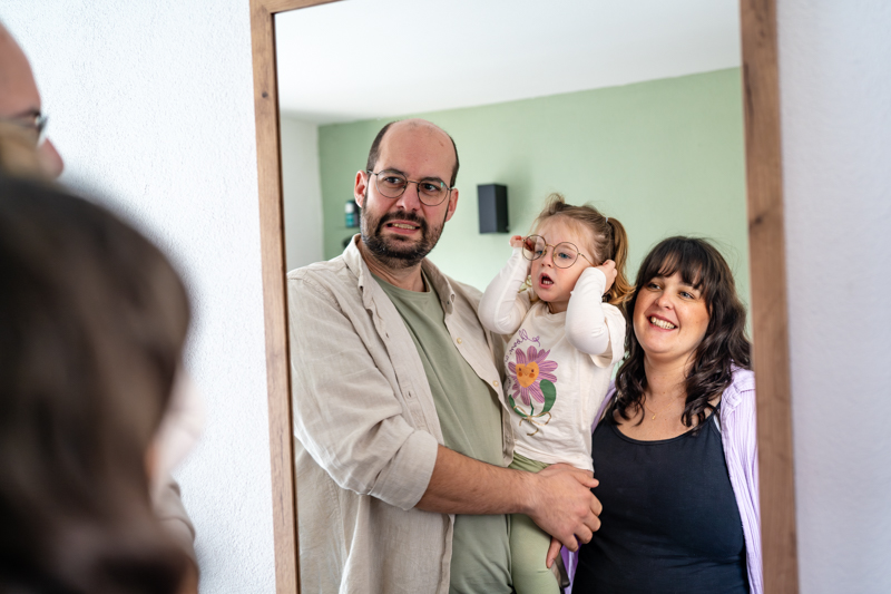 Vater hat Kleinkind auf dem Arm und lächelt in den Spiegel. Mutter steht mit dabei.