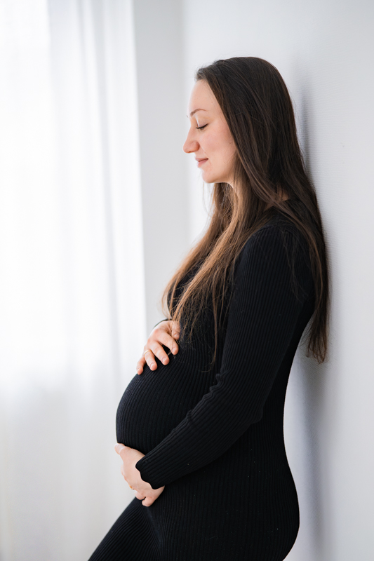 Schwangere Frau lehnt an der Wand und hält ihren Babybauch.